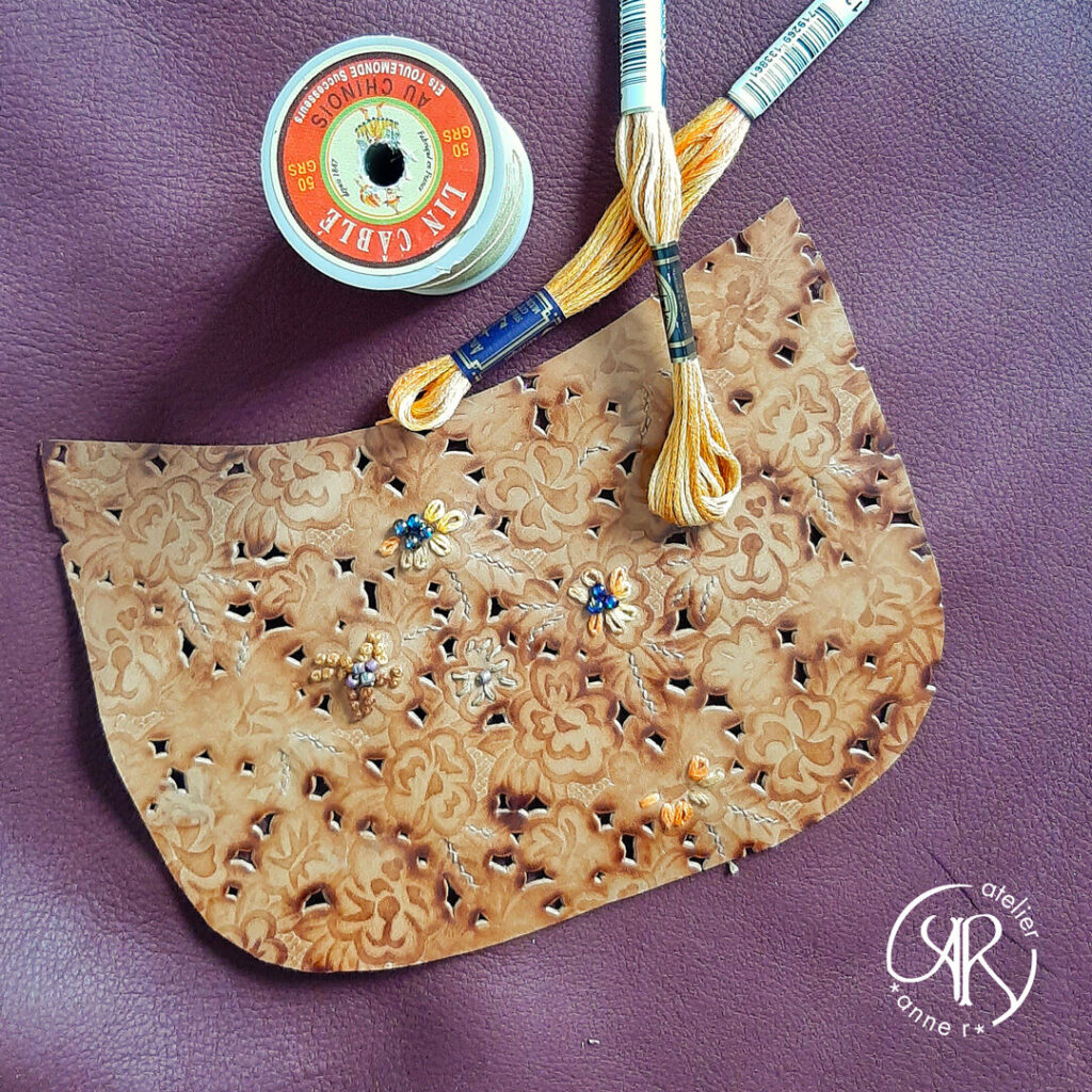 Photo d'un rabat de sac où l'on voit la broderie en cours. Le cuir est gaufré de fleurs et perforé autour de certaines fleurs. De couleur marron et glacé