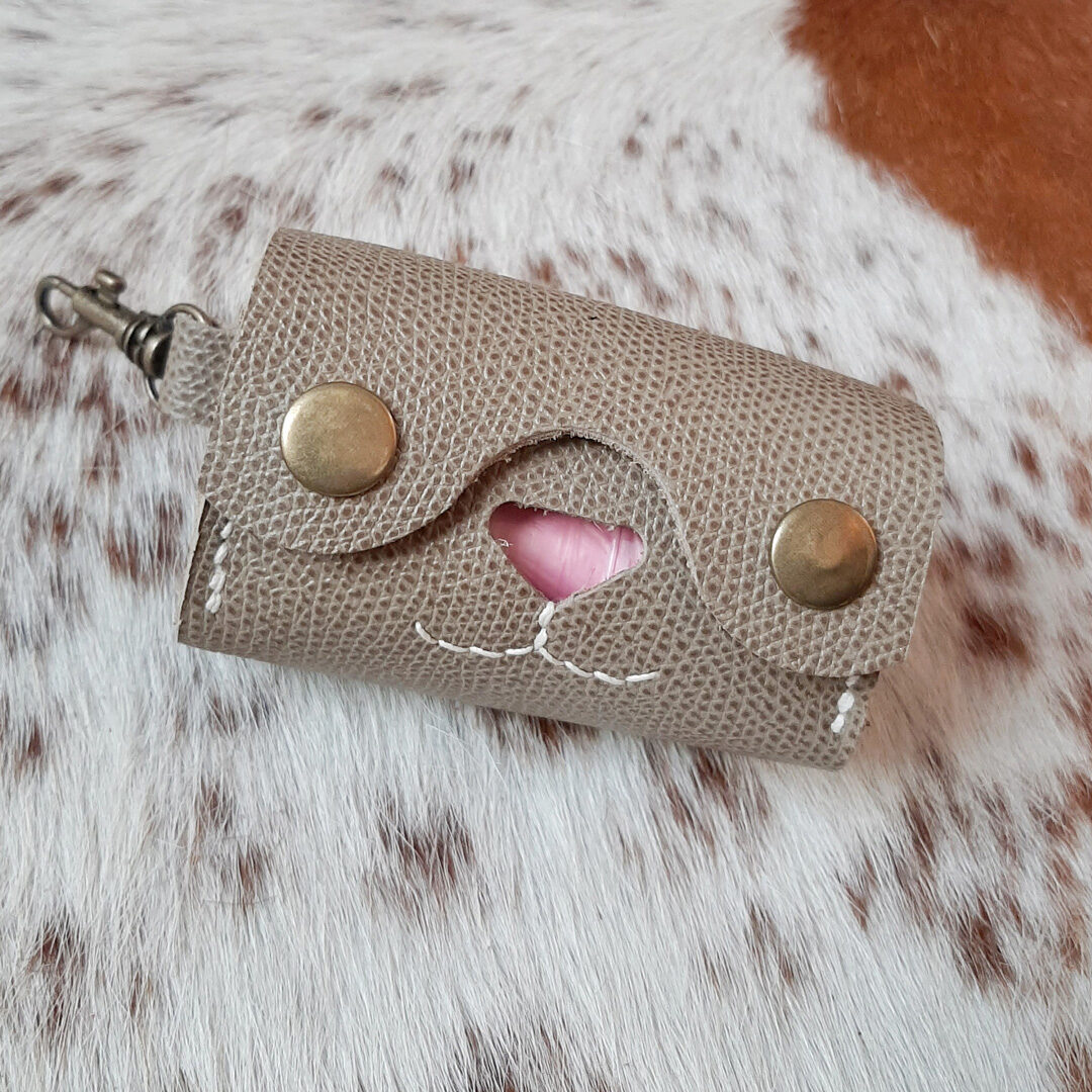 Un porte-sac à crottes élégant en cuir veau Epsom gris beige, avec une découpe en forme de cœur révélant un sac en plastique. Le design est épuré, avec deux boutons pression noirs et des coutures apparentes. Un mousqueton en métal suggère une fixation facile à une laisse ou un sac. L'ensemble est présenté sur un fond de peau de vache naturelle, soulignant le côté artisanal du produit.