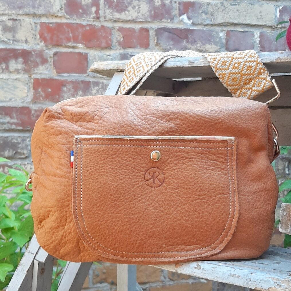 Sac bandoulière en cuir couleur caramel avec poche extérieure et une bandoulière sangle tissée à motifs géométriques, posé sur un escabeau en bois devant un mur de briques.