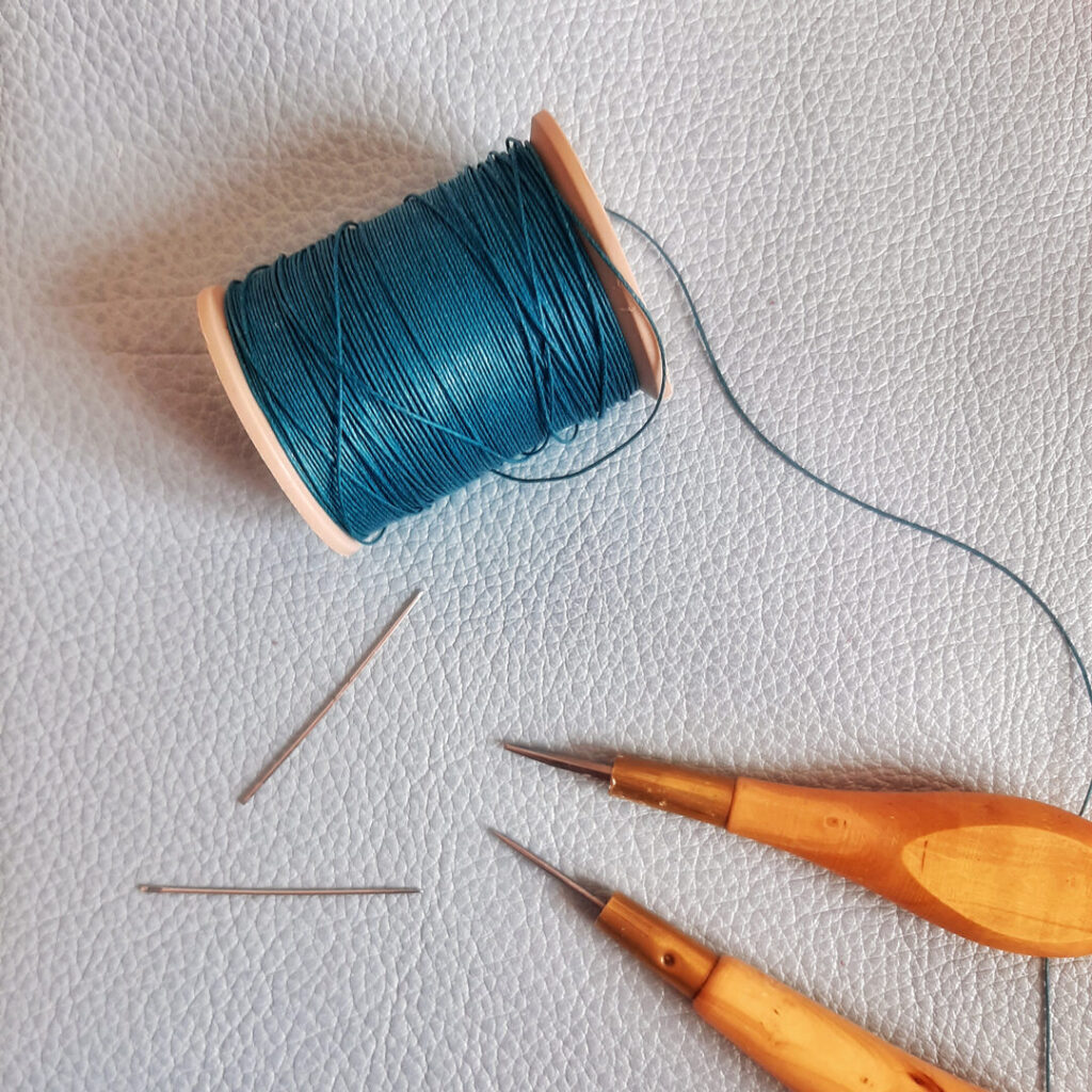 Bobine de fil de lin couleur bleu canard. posée sur un cuir grainé et accompagnée des outils de sellier maroquinier