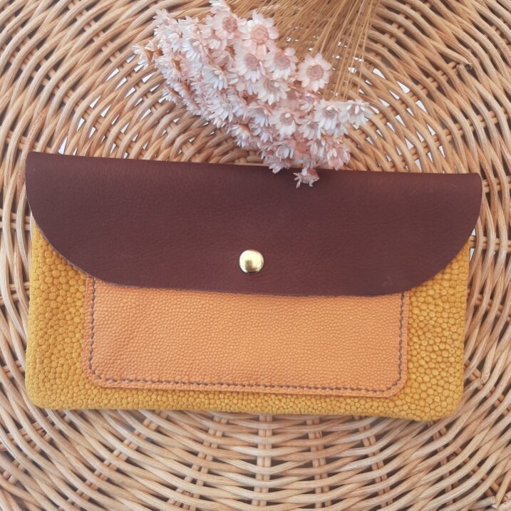 Pochette artisanale en cuir avec rabat bordeaux, poche extérieure en cuir fantaisie irisé grain caviar orange et corps en cuir fantaisie velours grain imitation galuchat camel, fermée par un bouton pression doré, posée sur un fond en rotin