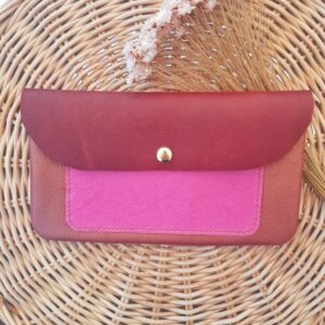 Pochette artisanale en cuir avec rabat bordeaux tannage végétal, poche extérieure cousue à la main, en cuir fantaisie imitation griffé rose fuchsia et corps en cuir fantaisie irisé grain caviar de couleur rouille, fermée par un bouton pression doré, posée sur un fond en rotin