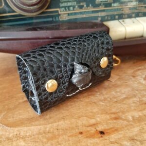 Etui pour rouleau de sachets de sacs à excréments. Cuir imitation alligator noir vernis. Deux boutons pressions dorés, un nez de forme d'un triangle évidé pour laisser passer les sachets en plastique, et une couture cousu main en lin blanc en dessous du nez qui représente les babines d'un chien ou chat. Sur le côté un mousqueton pour l'accrocher où vous le souhaitez.