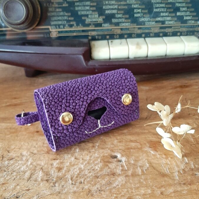 Etui pour rouleau de sachets de sacs à excréments. Cuir imitation grain galuchat toucher velours de couleur violet. Deux boutons pressions dorés, un nez de forme d'un triangle évidé pour laisser passer les sachets en plastique, et une couture cousu main en lin blanc en dessous du nez qui représente les babines d'un chien ou chat. Sur le côté un mousqueton pour l'accrocher où vous le souhaitez.