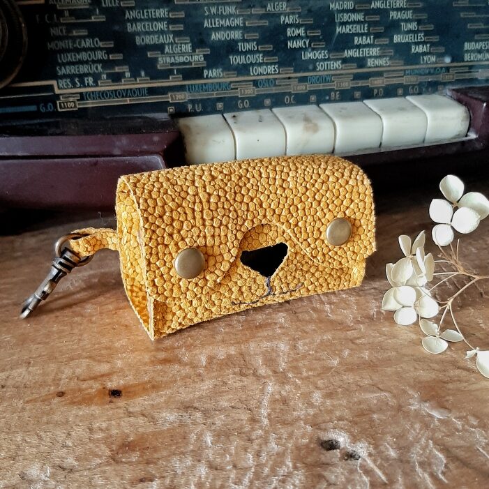 Etui pour rouleau de sachets de sacs à excréments. Cuir imitation grain galuchat toucher velours de couleur ocre. Deux boutons pressions laiton vieilli, un nez de forme d'un triangle évidé pour laisser passer les sachets en plastique, et une couture cousu main en lin marron en dessous du nez qui représente les babines d'un chien ou chat. Sur le côté un mousqueton pour l'accrocher où vous le souhaitez.