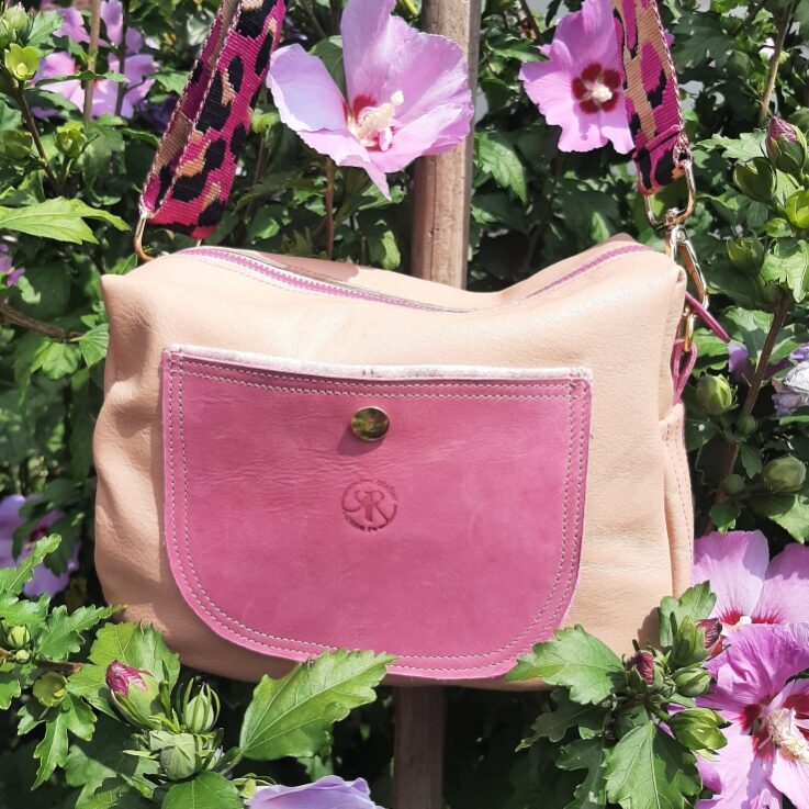 Sac bandoulière en cuir souple beige et une poche plaquée en cuir tannage végétal rose, se fermant par un bouton magnétique doré. Coutures contrastantes. fermeture à glissière rose assortie à la poche. Bandoulière tissées fuchsia et noir motif pelage panthère.