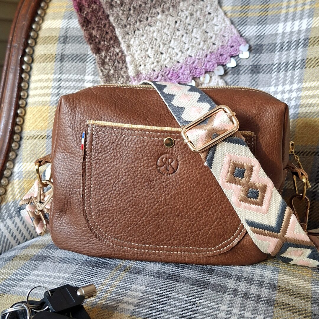 Sac bandoulière cuir pleine fleur grainé marron. Sac posé sur un fauteuil . Poche plate cousue en double surpiqûre sur le devant, et se fermant par un bouton pression doré, petit drapeau bleu, blanc, rouge cousu sur le côté de la poche.. Bandoulière tissé géométrique dans les tons rose, gris, marron et blanc cassé. Elle est réglable et amovible