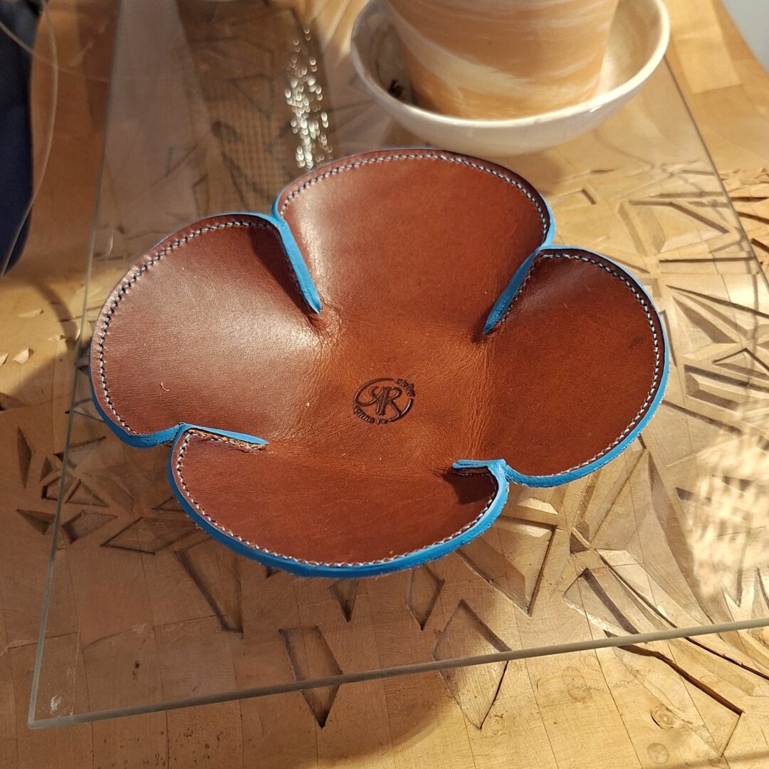 Vide poche en cuir tannage végétal de couleur marron . Couture réalisée à la main au point sellier avec un fil de couleur turquoise. Teinture des tranches en bleu turquoise. Logo estampé dans le fond du vide-poches.