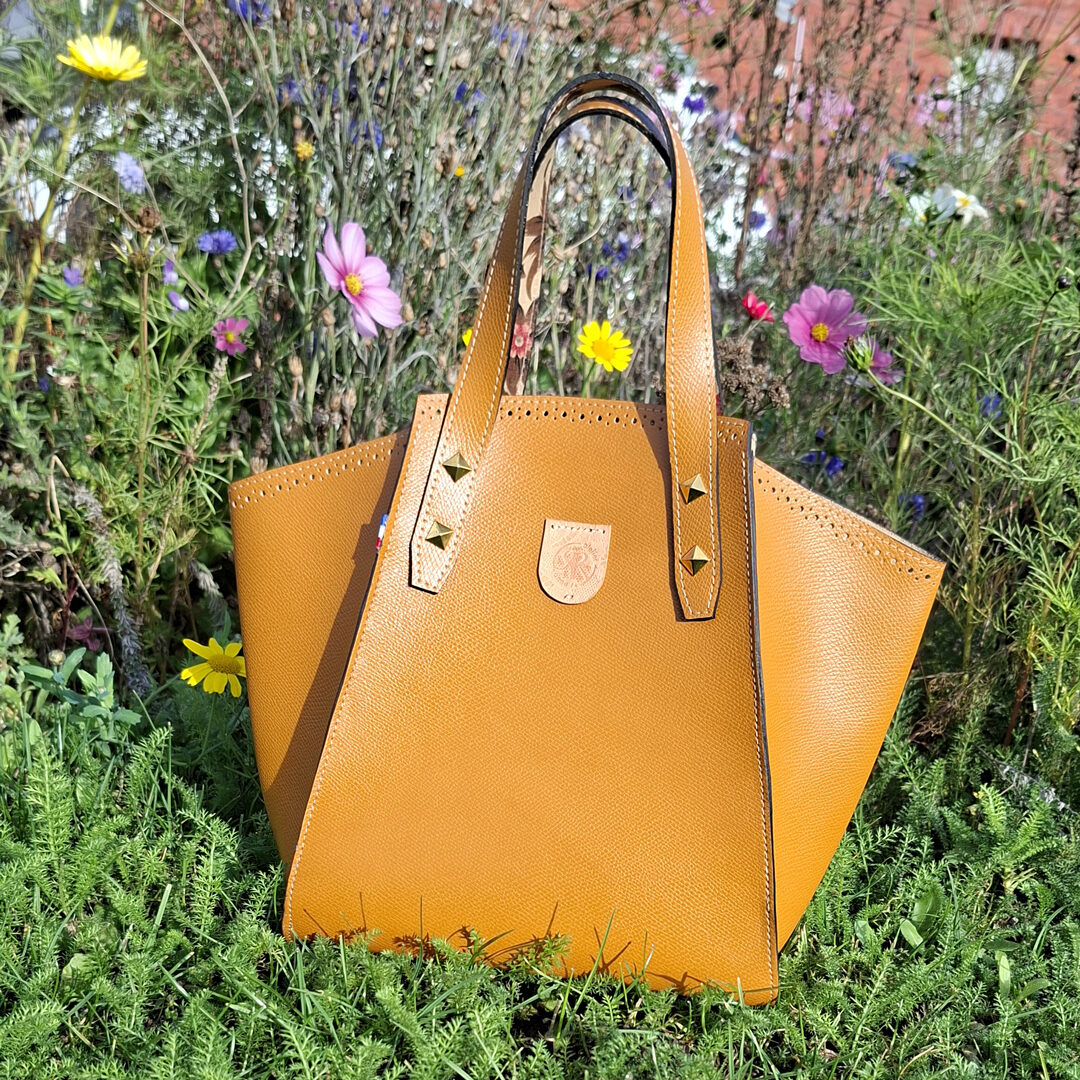 Sac cabas en cuir epsom, de marron ocre, couture finition main, avec soufflets souples repliables, posé dans des fleurs. Des rivets pyramidales fixés sur le bas des poignées. Finition des tranches en noir. création artisanale française Atelier Anne R
