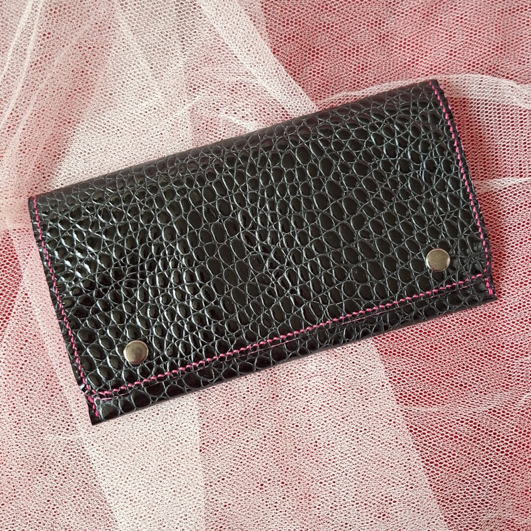 Compagnon en cuir vernis noir, gaufrage imitation crocodile. Deux boutons pressions métalliques argenté sur le rabat permettent de bien fermer cette pochette compagnon. Couture main au point sellier au fil de lin couleur fuchsia.