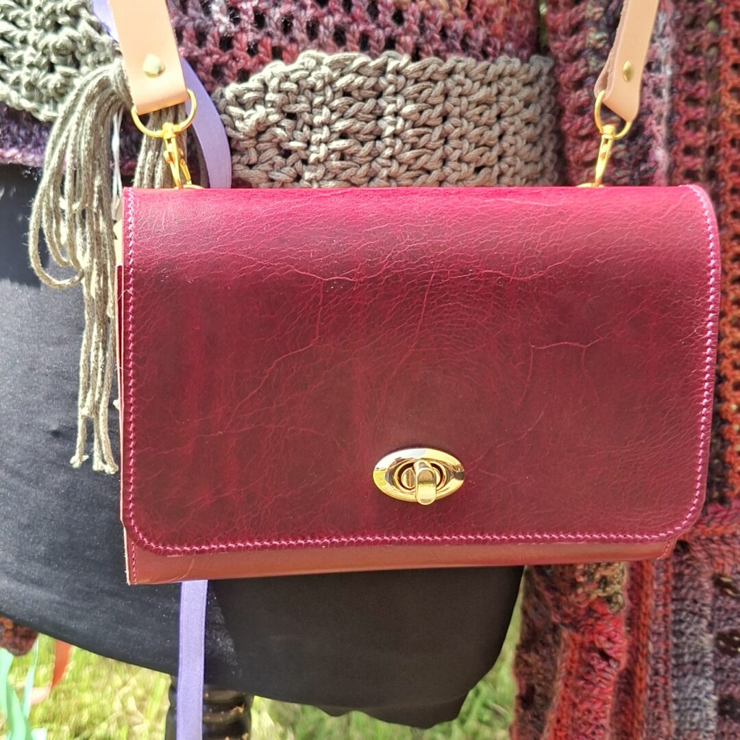 Sac bandoulière artisanal en cuir pleine fleur bordeaux porté à l’épaule avec fermeture métallique dorée.