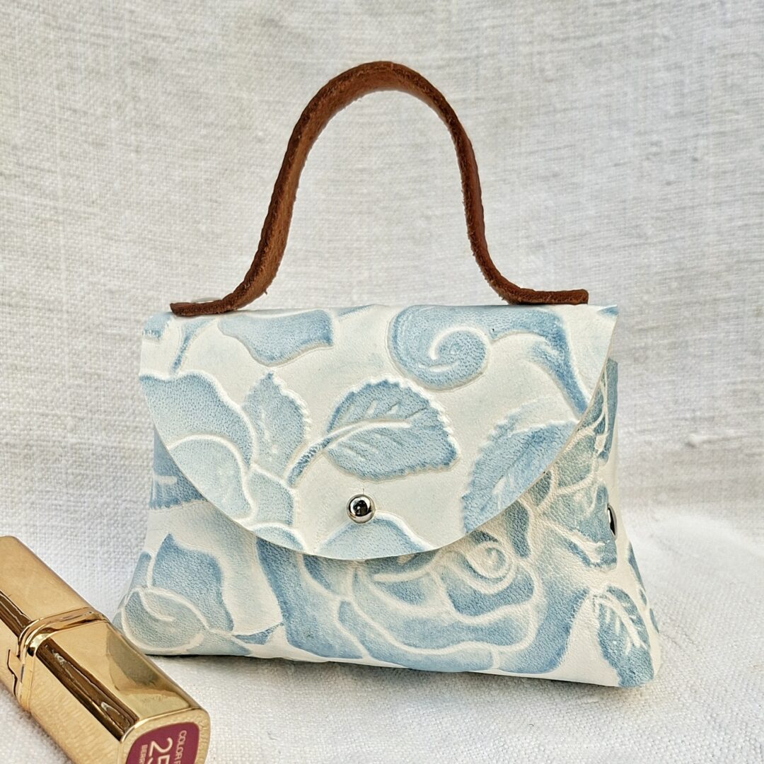 Petit sac en cuir de chèvre embossé à fleurs bleu clair sur fond blanc avec poignée en cuir marron pleine fleur.