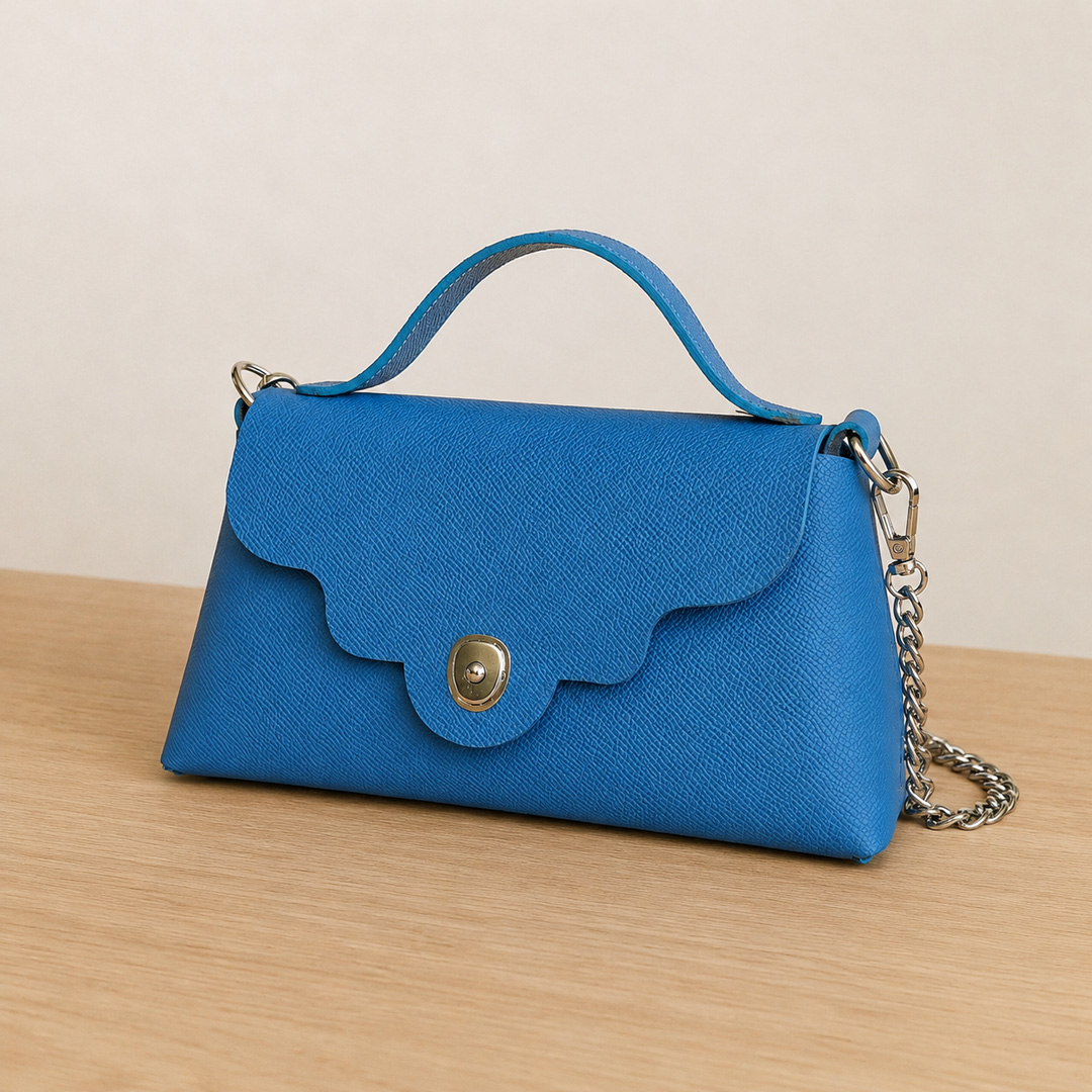 Sac bandoulière Capucine en cuir Epsom bleu présenté sur un meuble en bois avec fond neutre, fabrication artisanale française.